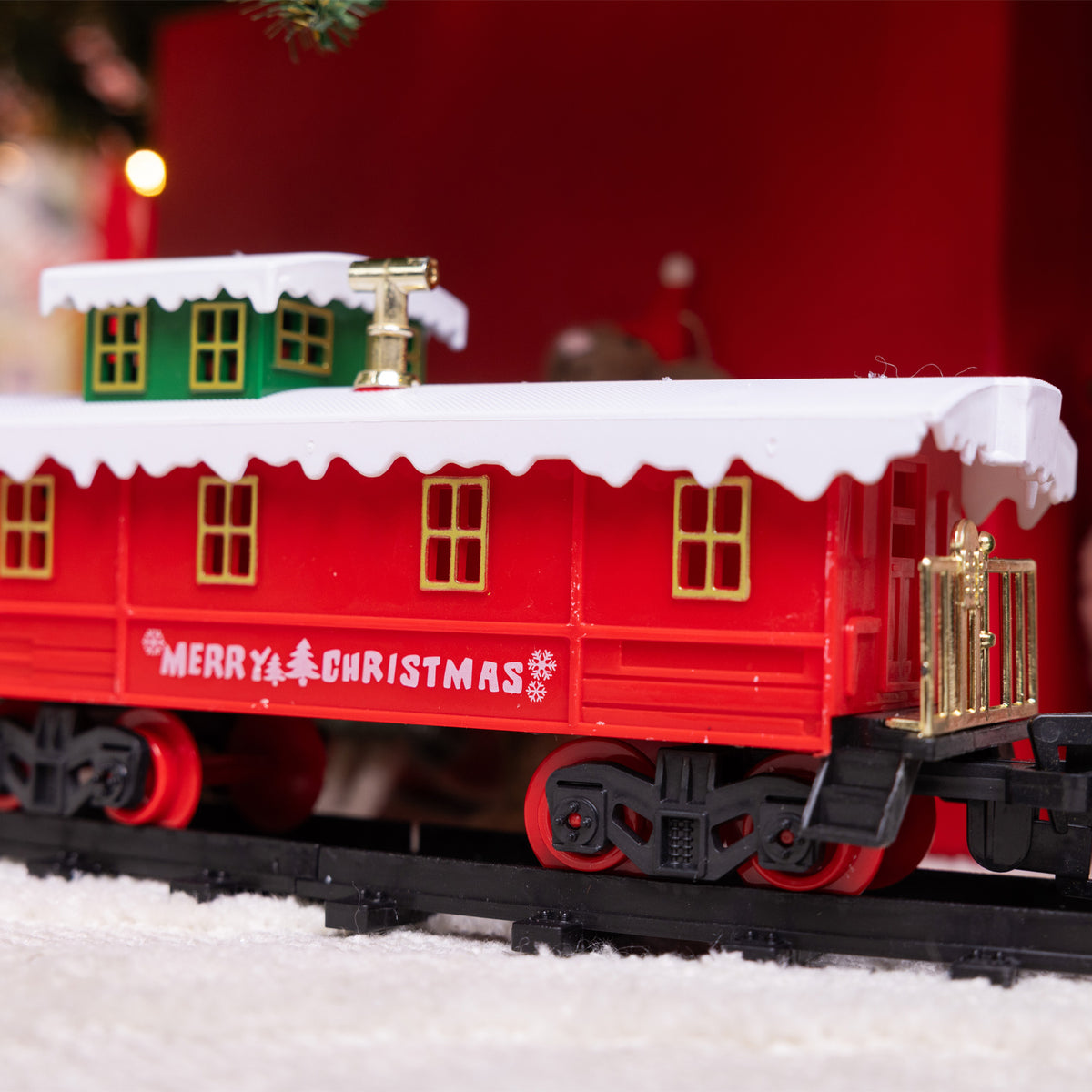 A red Christmas-themed train car on a black track, decorated with white snow, gold window frames and trim, and 'MERRY CHRISTMAS' written on the side, set against a festive red and dark background.
