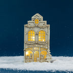 Completed wooden Christmas shop puzzle displayed with its warm LED lights glowing through the windows. Features intricate festive detailing, gift displays, decorative stars and a Christmas tree, styled amongst faux snow.