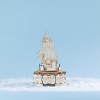 Completed wooden Santa music box featuring Santa beside a decorated Christmas tree and surrounded by stacked presents, all set on an intricately detailed festive base. Styled amongst faux snow against a soft blue backdrop.