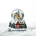A charming Christmas snow globe featuring a snowman among snowy trees, set above a warmly lit festive cottage surrounded by snow.