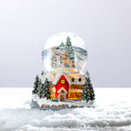 A charming Christmas snow globe featuring a snowman among snowy trees, set above a warmly lit festive cottage surrounded by snow.