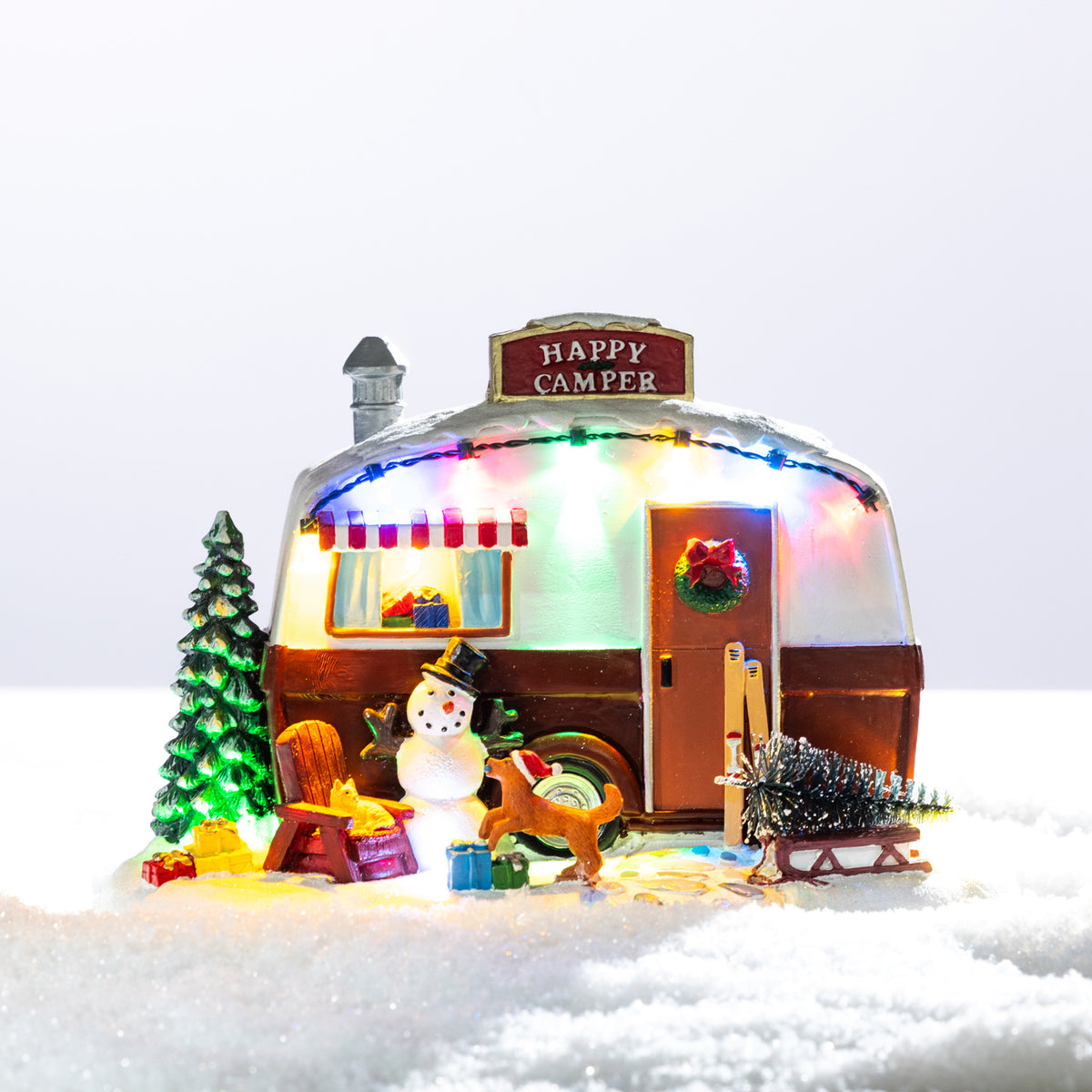 A whimsical illuminated Christmas camper scene featuring a snowman, reindeer, festive tree and colourful lights nestled in a snowy setting.