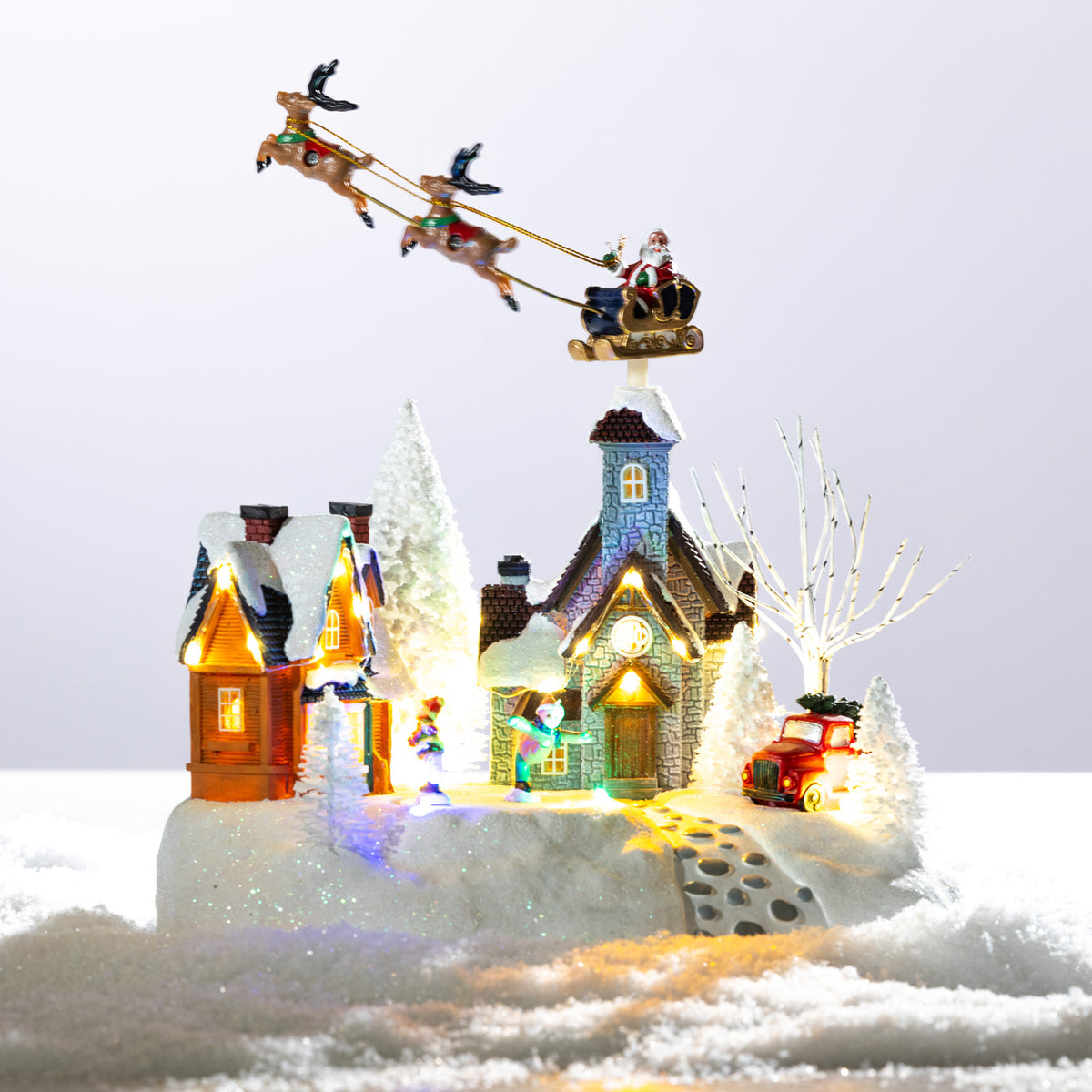 A whimsical illuminated Christmas village featuring snowy cottages, festive figurines, a red car and Santa flying overhead in his reindeer-pulled sleigh.