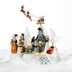 A whimsical illuminated Christmas village featuring snowy cottages, festive figurines, a red car and Santa flying overhead in his reindeer-pulled sleigh.