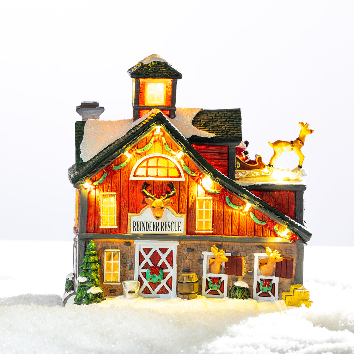A whimsical illuminated Reindeer Rescue barn featuring festive garlands, cosy glowing windows and adorable reindeer nestled in a snowy setting.