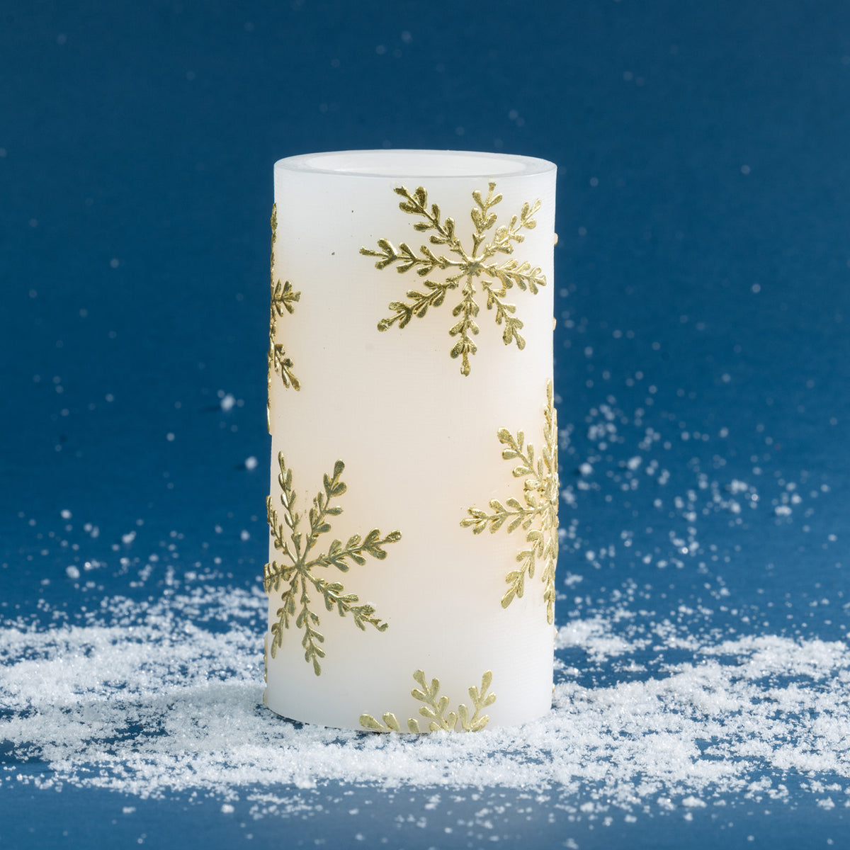 Large white LED candle featuring an all-over metallic gold snowflake design. Styled against a rich blue background with scattered faux snow, highlighting its taller decorative shape.