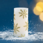 Large white LED Christmas candle with metallic gold snowflake detailing, styled with soft golden bokeh lighting and faux snow to create a warm seasonal display.
