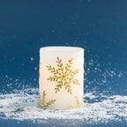 Small white LED candle decorated with elegant metallic gold snowflake detailing. Styled against a deep blue festive backdrop with scattered faux snow and a soft warm glow.