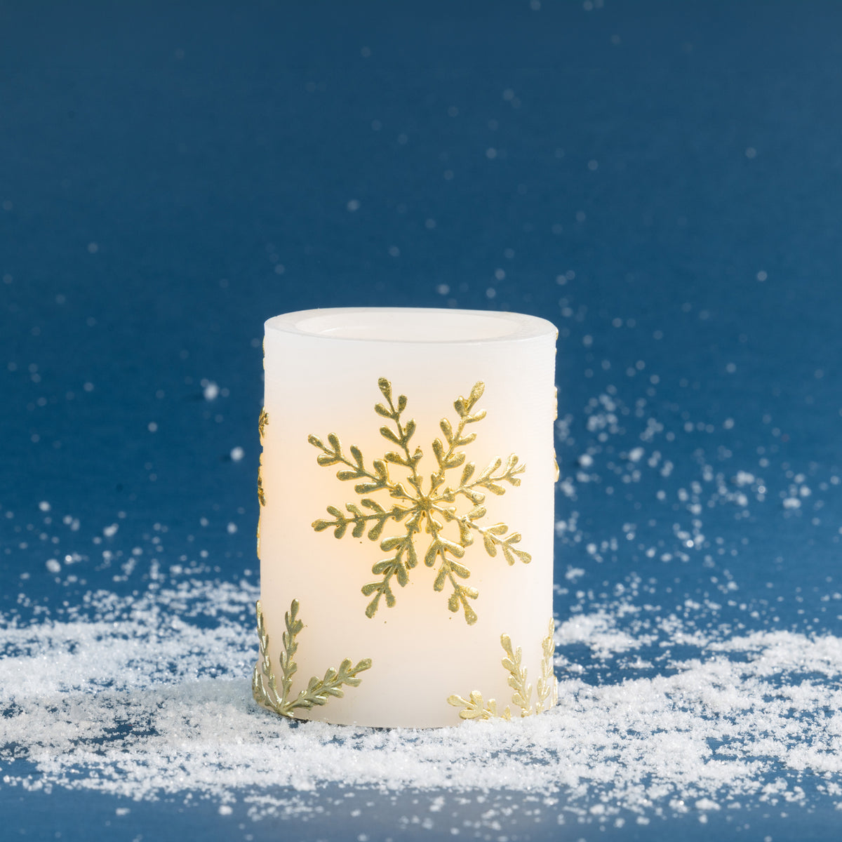 Small white LED candle decorated with elegant metallic gold snowflake detailing. Styled against a deep blue festive backdrop with scattered faux snow and a soft warm glow.