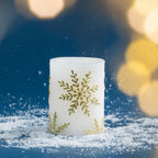 Small white LED Christmas candle featuring gold snowflake motifs, photographed with warm golden bokeh lighting and scattered faux snow for a cosy festive look.