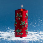 Large red pillar candle featuring raised green holly leaves and berry accents for a classic Christmas look. Shown lit against a deep blue backdrop with scattered faux snow.