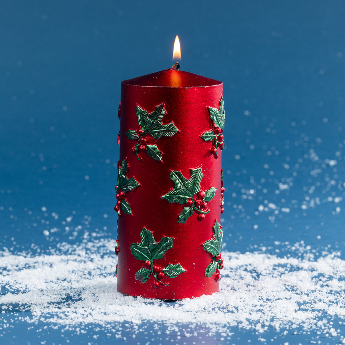 Large red pillar candle featuring raised green holly leaves and berry accents for a classic Christmas look. Shown lit against a deep blue backdrop with scattered faux snow.