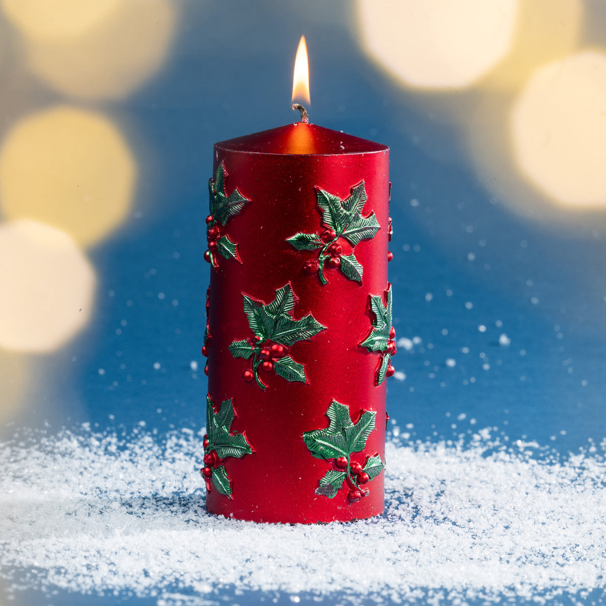 Large red holly and berry Christmas candle shown burning amongst faux snow, with soft golden bokeh lighting creating a warm seasonal setting.