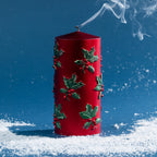 Large red pillar candle decorated with raised green holly leaves and berries, photographed after extinguishing with curling smoke above the wick.