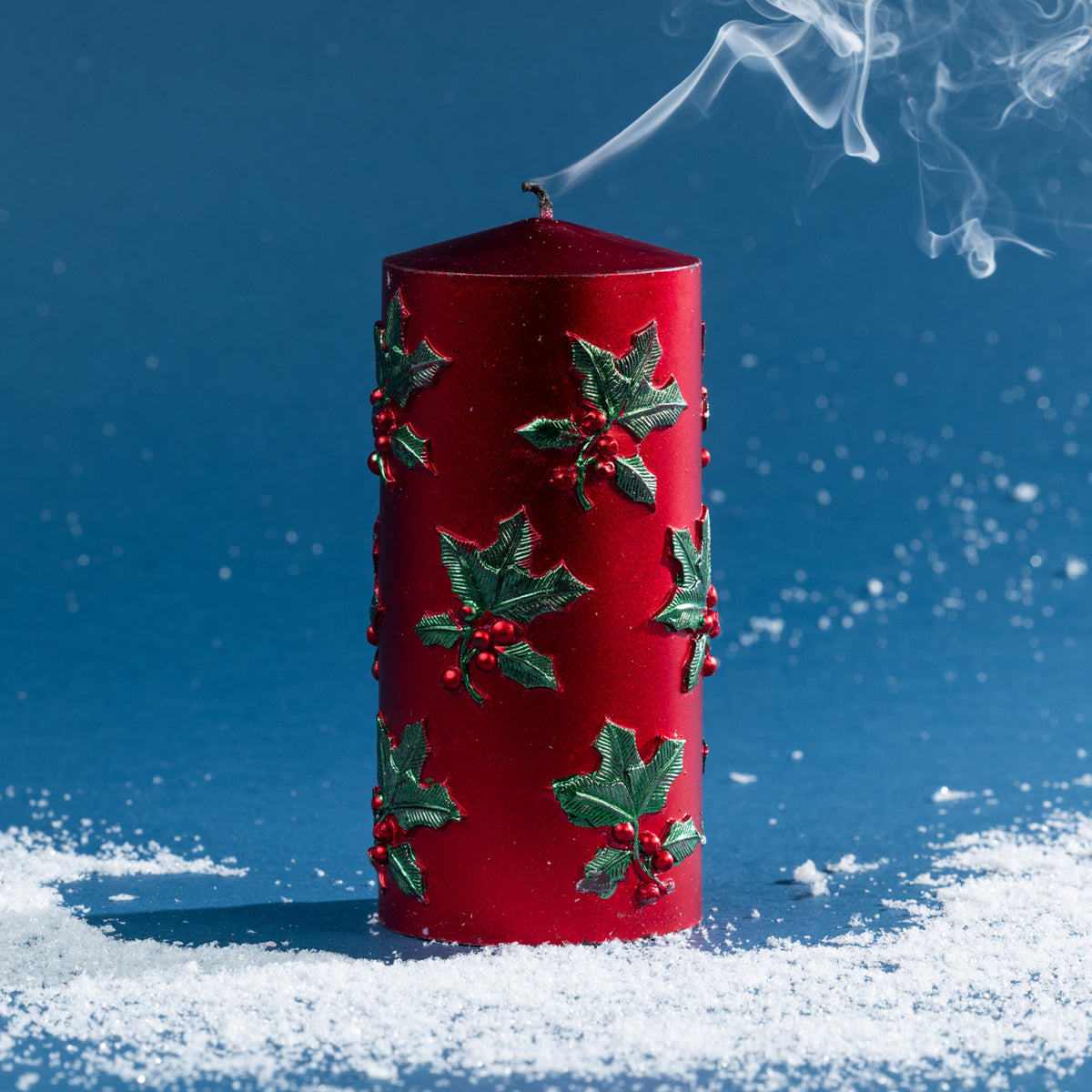 Large red pillar candle decorated with raised green holly leaves and berries, photographed after extinguishing with curling smoke above the wick.