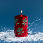 Small red pillar candle decorated with raised green holly leaves and red berry detailing. Shown burning against a rich blue festive backdrop with scattered faux snow.