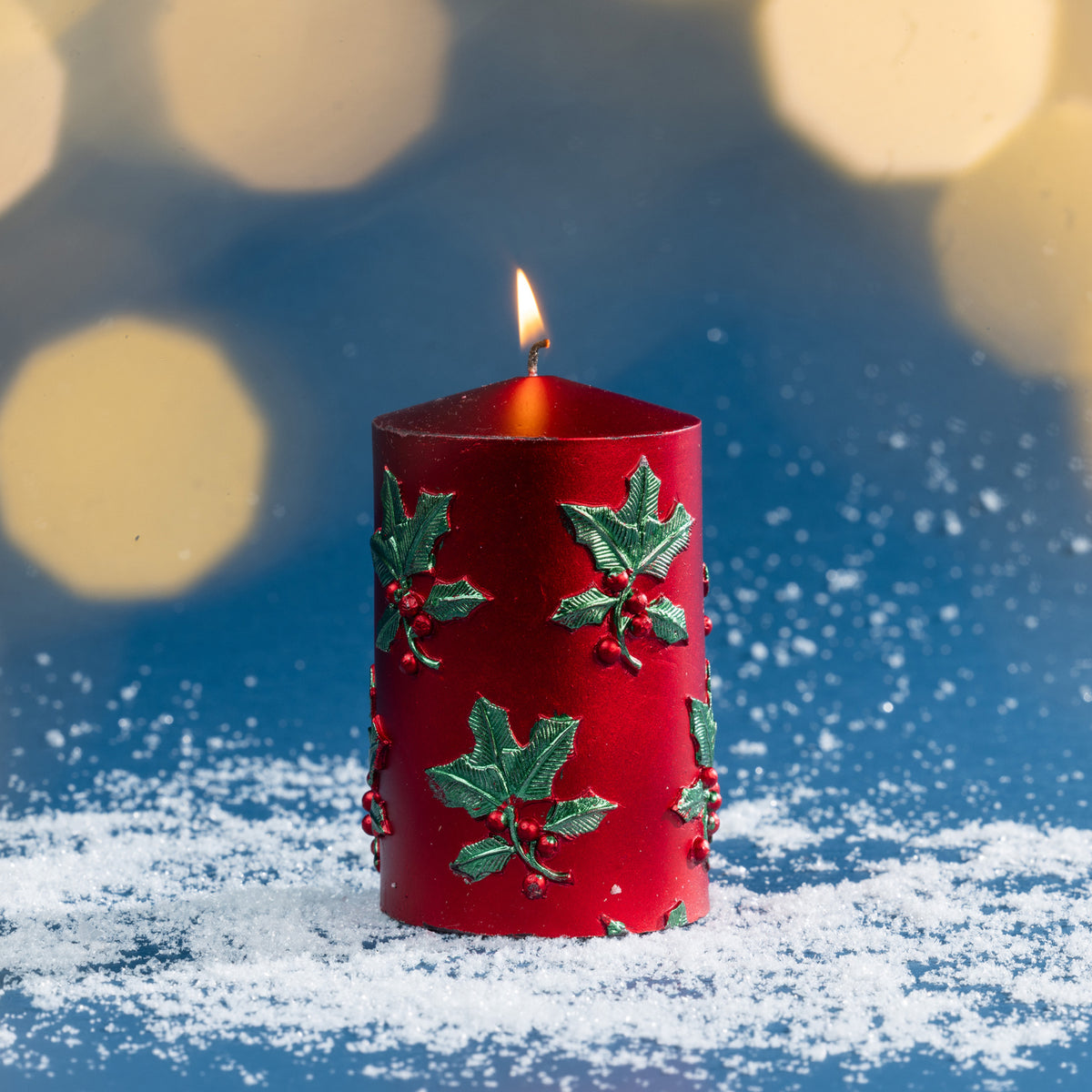 Small red holly and berry Christmas candle photographed burning amongst faux snow, with warm golden bokeh lights adding a cosy festive glow.