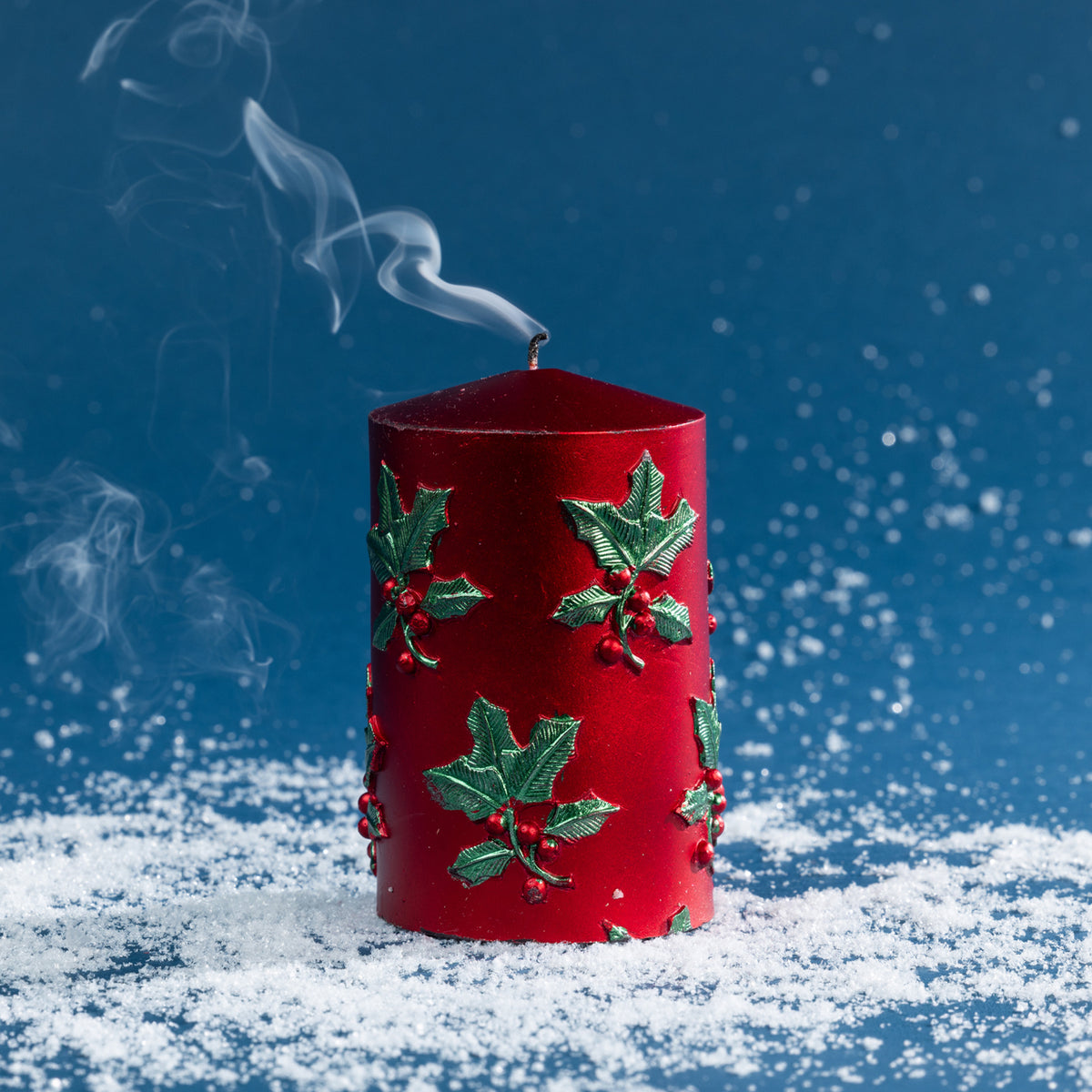 Small red pillar candle with raised holly and berry detailing shown after extinguishing, with smoke rising gently from the wick.
