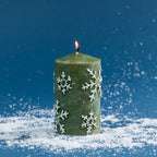 Green pillar candle decorated with raised white snowflake detailing and a lightly frosted finish. Photographed lit against a deep blue festive backdrop with scattered faux snow.