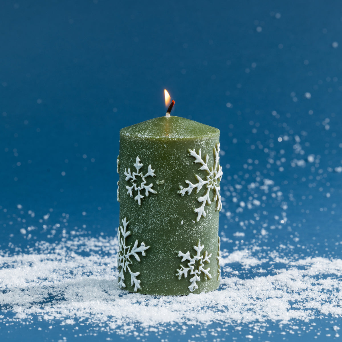 Green pillar candle decorated with raised white snowflake detailing and a lightly frosted finish. Photographed lit against a deep blue festive backdrop with scattered faux snow.