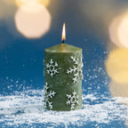 Green snowflake pillar candle shown burning amongst faux snow, with warm golden bokeh lighting creating a cosy Christmas atmosphere.