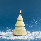 Christmas tree-shaped candle finished in shimmering gold with textured tiered branches. Shown lit against a deep blue festive backdrop with scattered faux snow.
