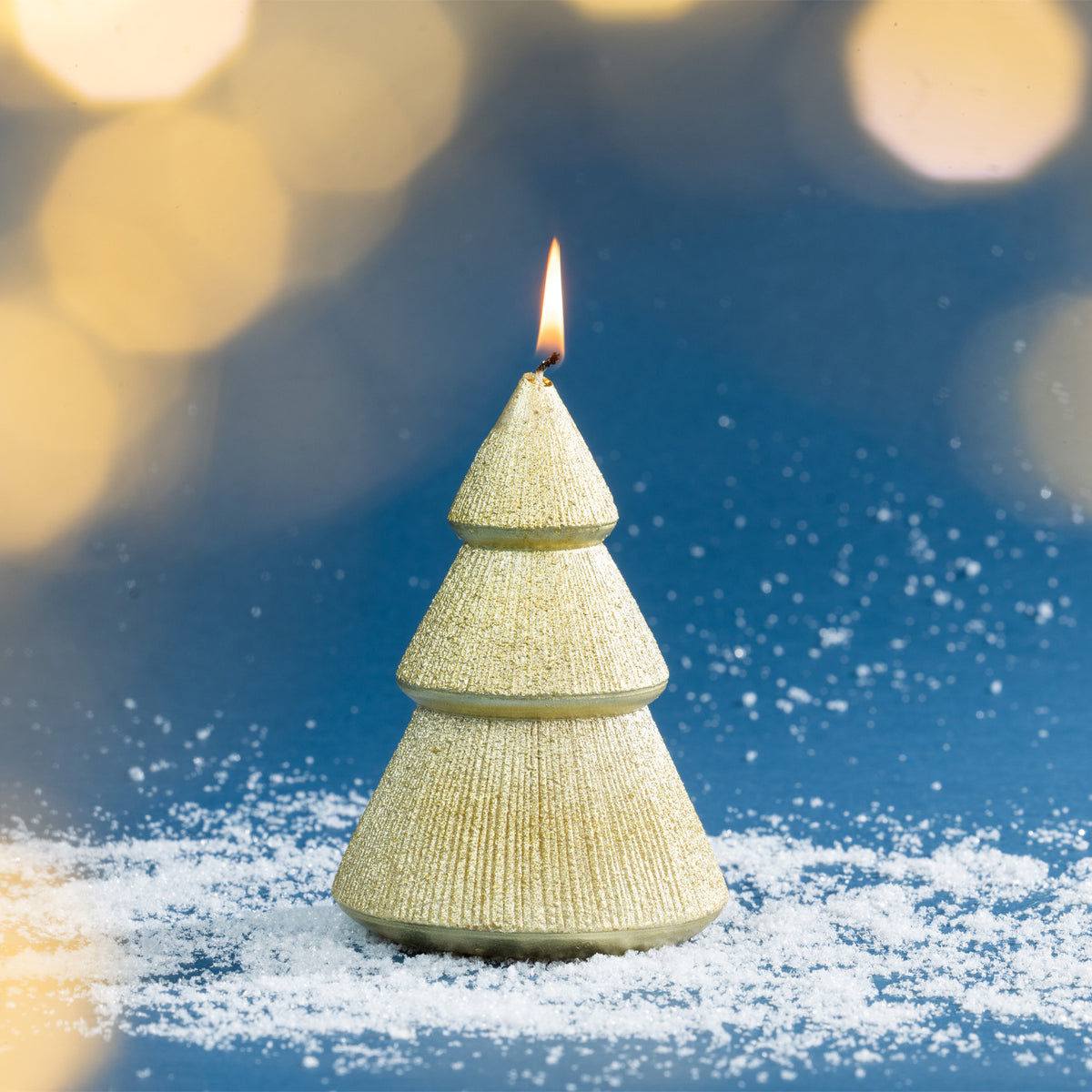 Gold Christmas tree candle photographed burning amongst faux snow, complemented by warm golden bokeh lights for an elegant festive look.