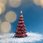 Metallic red Christmas tree candle shown burning against a blue backdrop with soft golden bokeh lighting and scattered faux snow.