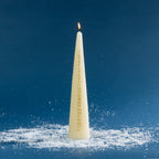 Tall off-white taper candle featuring gold numbers from 1 to 24 down the front, designed for counting down the days to Christmas. The candle is shown lit against a deep blue background with scattered faux snow for a festive winter setting.