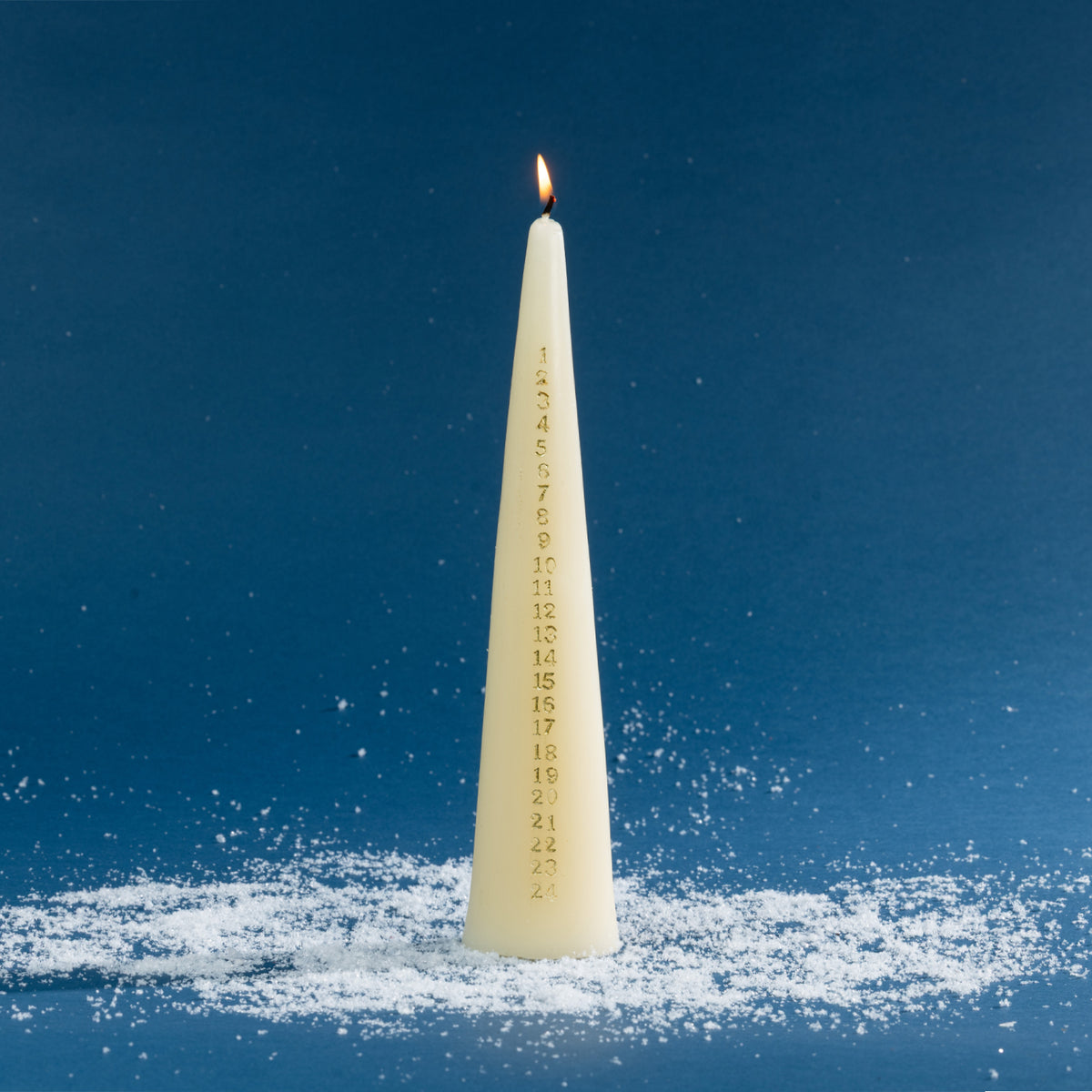 Tall off-white taper candle featuring gold numbers from 1 to 24 down the front, designed for counting down the days to Christmas. The candle is shown lit against a deep blue background with scattered faux snow for a festive winter setting.