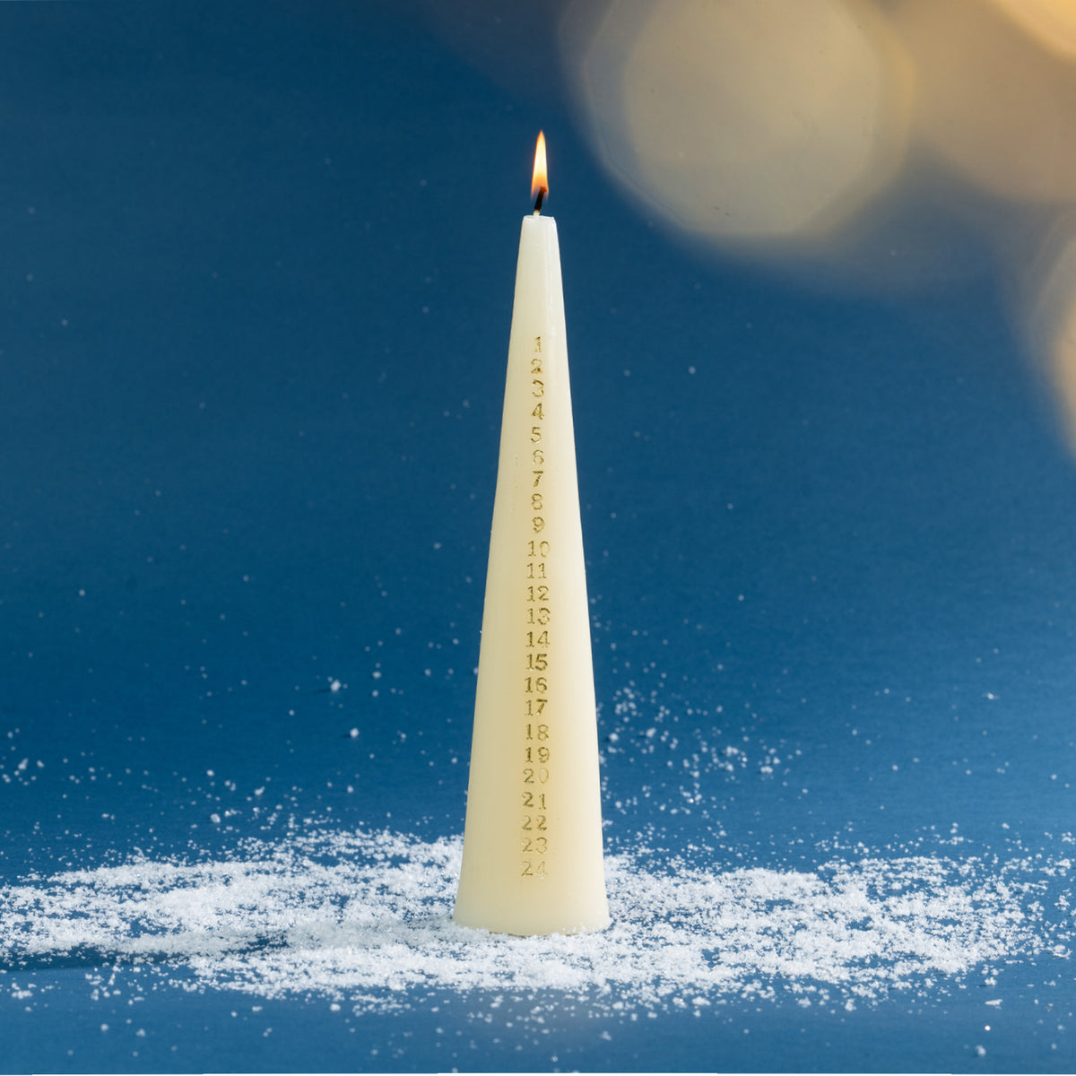 Off-white Christmas countdown taper candle with gold numbered markings from 1 to 24 and a glowing flame. Styled against a rich blue background with soft golden light effects and scattered faux snow.