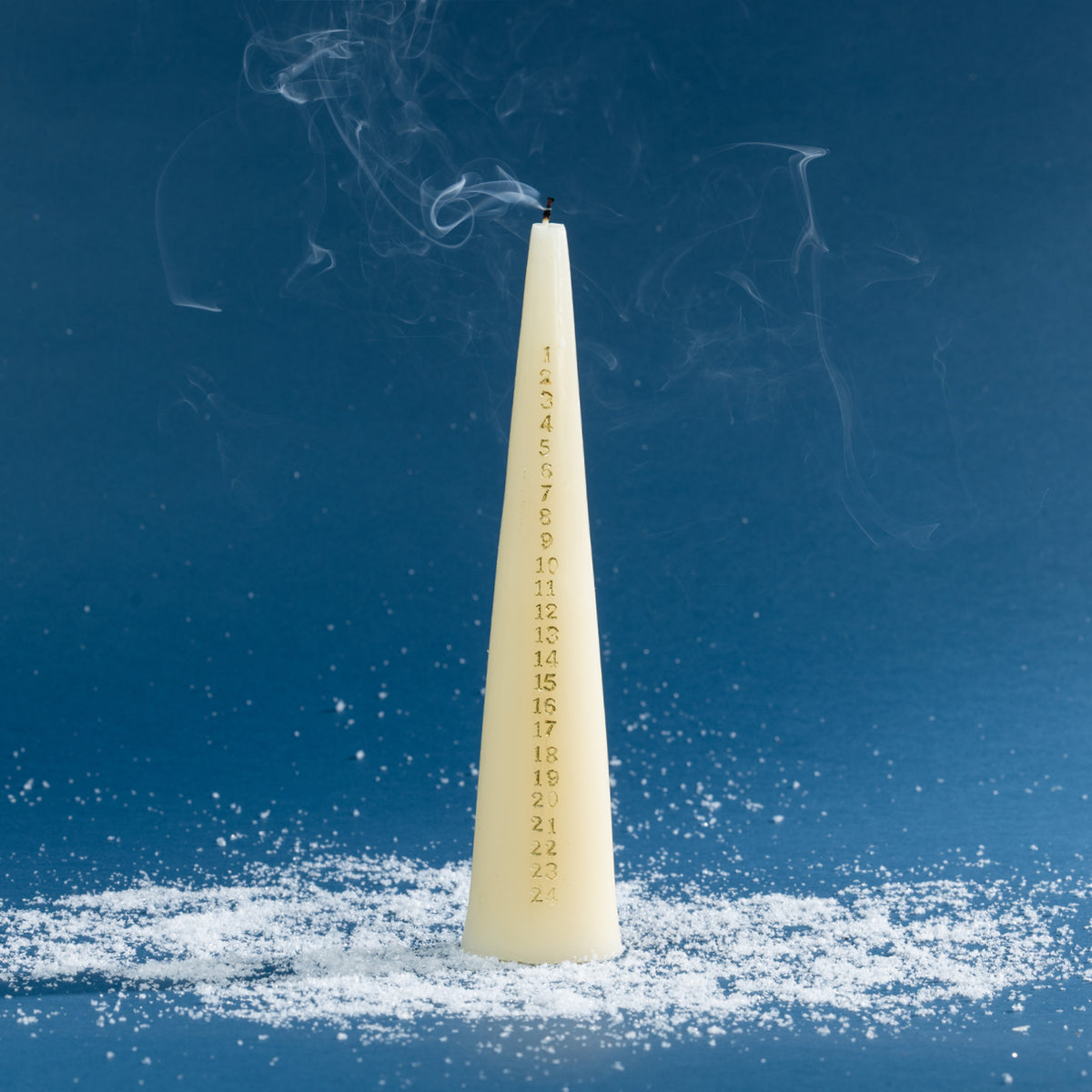 Off-white taper candle featuring gold countdown numbers from 1 to 24, shown just after the flame has been extinguished with wisps of smoke curling above the wick. Styled against a deep blue background with scattered faux snow.