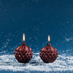 Set of two round bauble-shaped candles in a deep metallic red finish, featuring embossed geometric detailing and lit wicks.