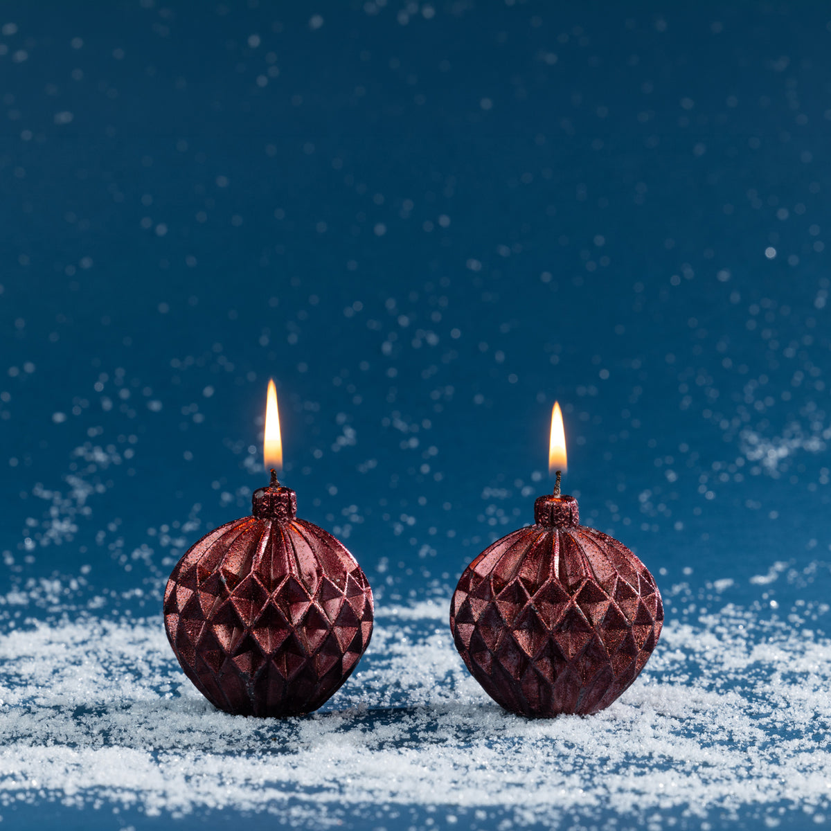 Set of two round bauble-shaped candles in a deep metallic red finish, featuring embossed geometric detailing and lit wicks.