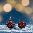 Pair of red bauble-shaped Christmas candles shown burning amongst faux snow with warm golden bokeh lights creating a festive atmosphere.