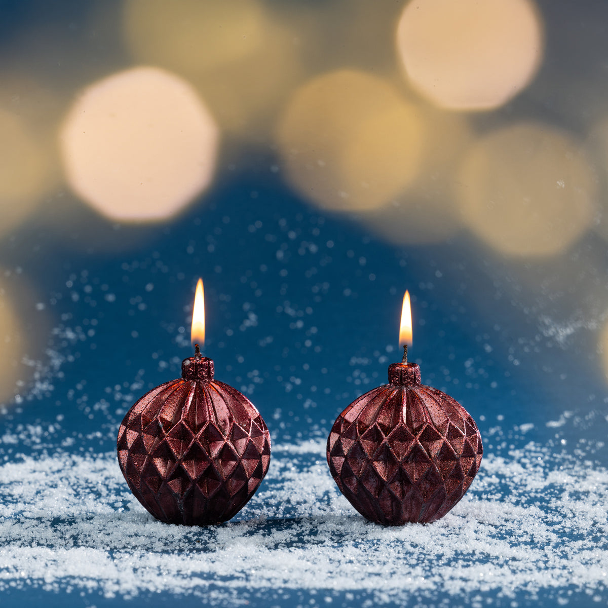 Pair of red bauble-shaped Christmas candles shown burning amongst faux snow with warm golden bokeh lights creating a festive atmosphere.
