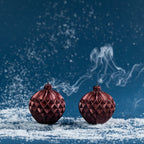 Pair of metallic red bauble candles shown after extinguishing, with curling smoke rising from the wicks against a rich blue Christmas backdrop.