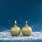 Set of two round bauble-shaped candles finished in shimmering gold, featuring elegant curved ribbed detailing and lit wicks. Styled against a deep blue festive backdrop with scattered faux snow.