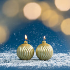 Pair of gold bauble-shaped Christmas candles shown burning amongst faux snow, with warm golden bokeh lighting creating a soft festive atmosphere.