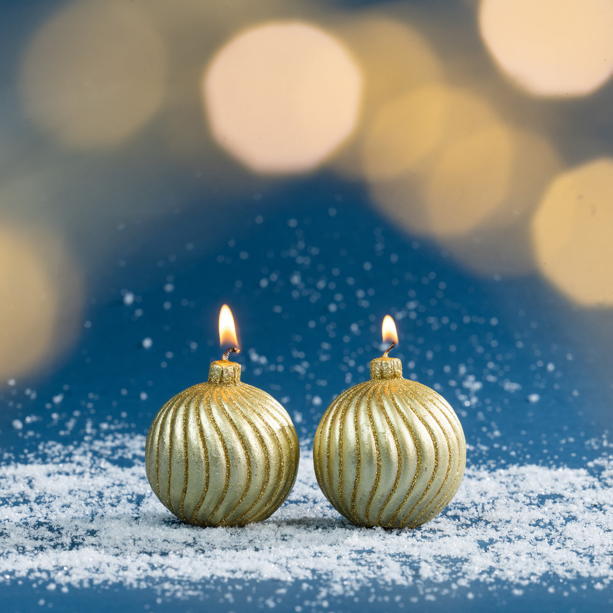 Pair of gold bauble-shaped Christmas candles shown burning amongst faux snow, with warm golden bokeh lighting creating a soft festive atmosphere.