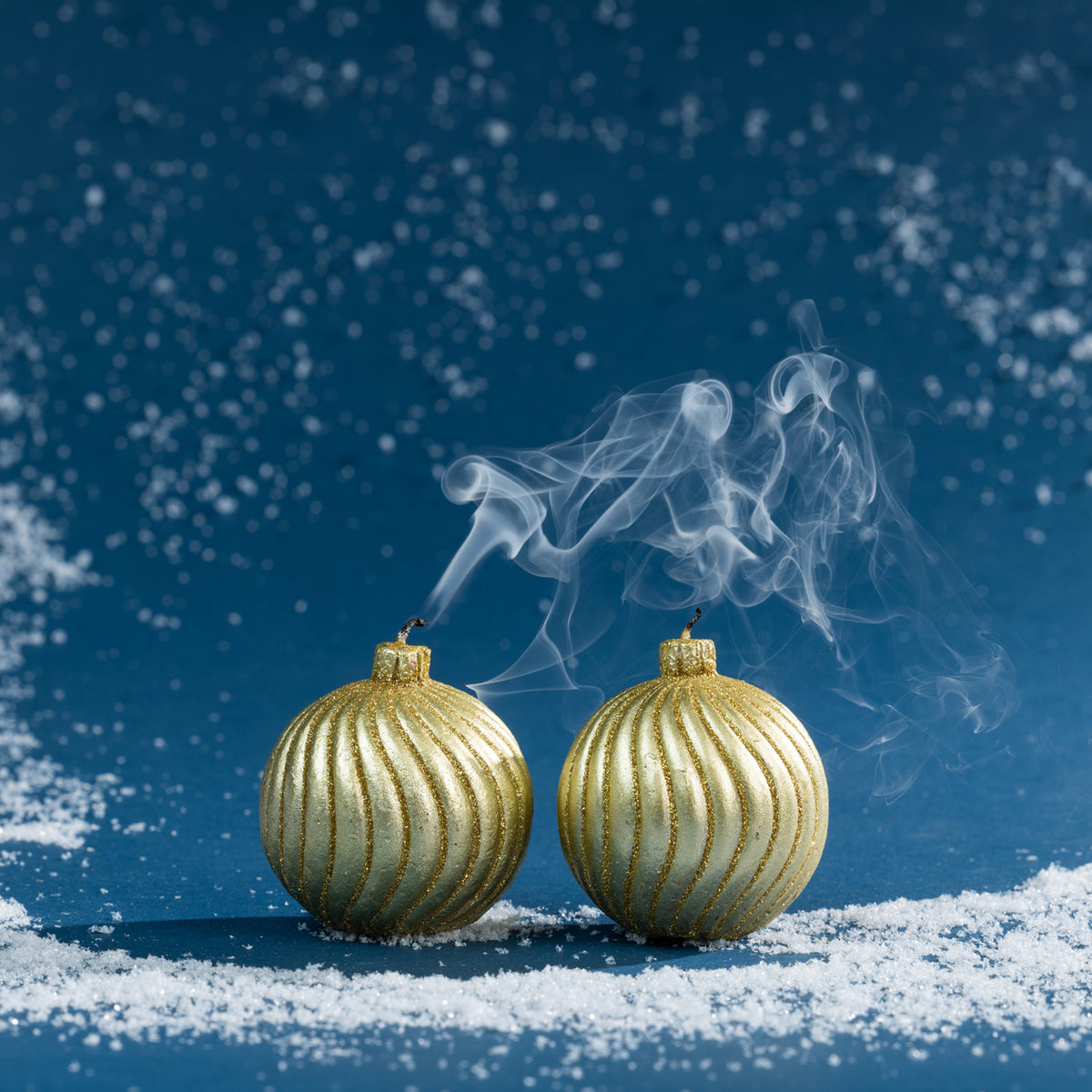 Pair of gold bauble candles shown after extinguishing, with delicate smoke curling from the wicks against a rich blue Christmas backdrop.