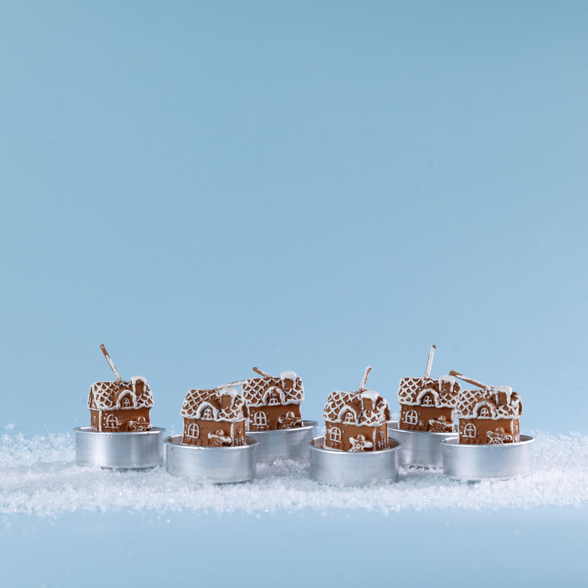 Set of six gingerbread house-shaped tealight candles featuring white icing-style detailing, snowy rooftops and charming miniature windows and doors. Styled together amongst faux snow.