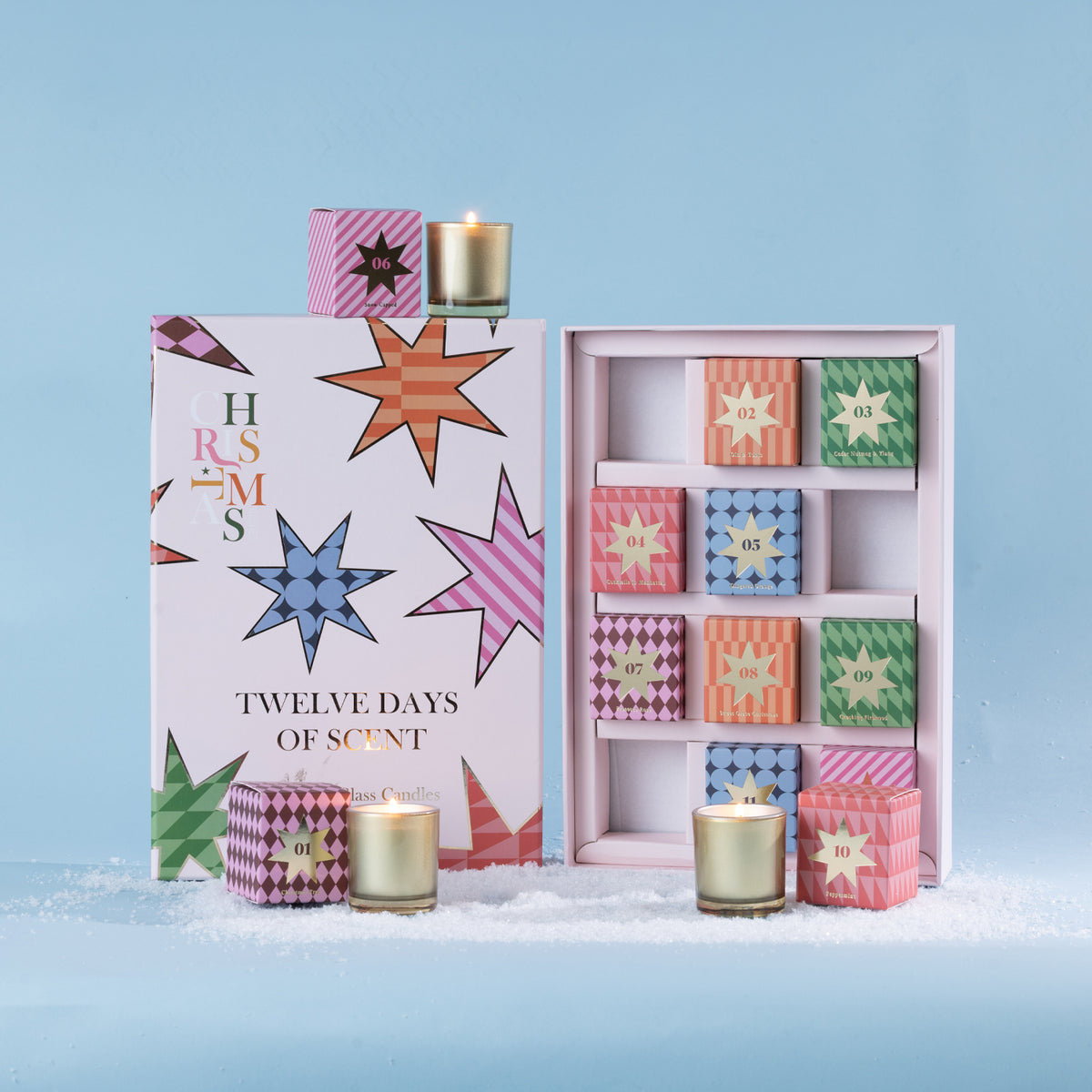 Christmas Wishes Twelve Days of Scent set featuring 12 individually boxed glass candles in assorted colourful festive designs, displayed with the open presentation box on a snowy light blue backdrop.