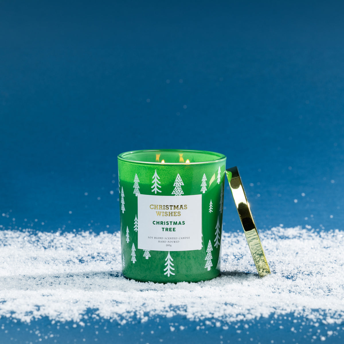 Christmas Wishes Christmas Tree 283g scented candle in a green glass vessel with white tree pattern, lit with a gold lid resting beside it on a snowy blue backdrop.