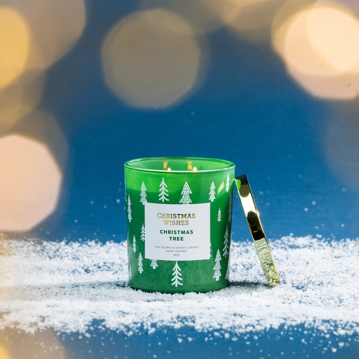 Christmas Wishes Christmas Tree 283g scented candle in green glass, lit with its gold lid beside it, displayed against a festive blue background with warm blurred lights.