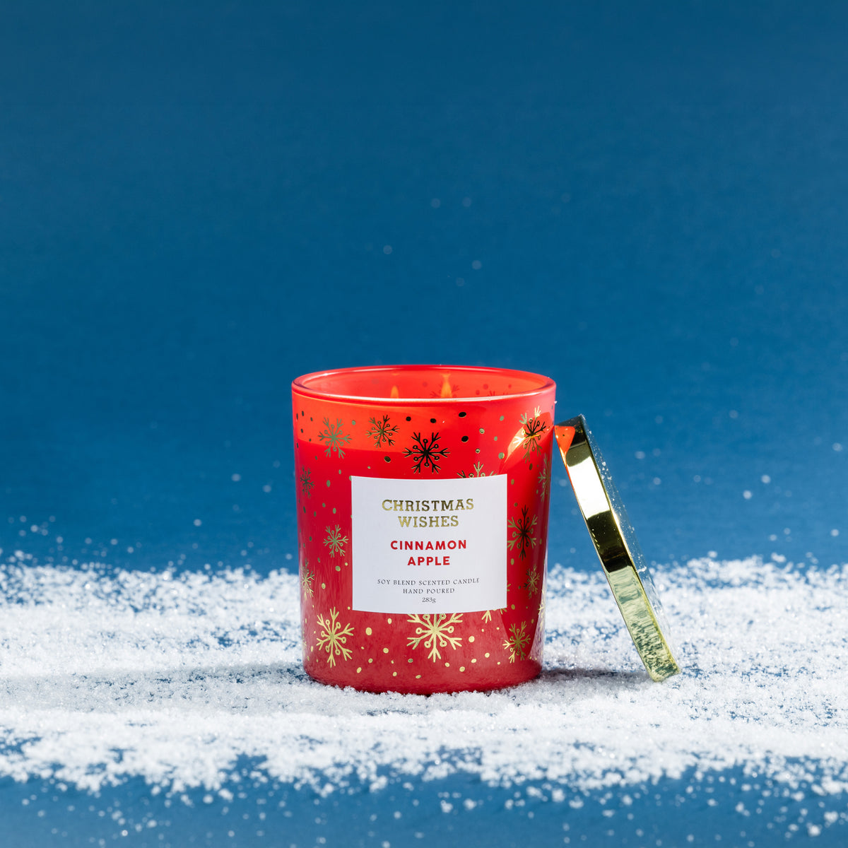 Christmas Wishes Cinnamon Apple 283g scented candle in a red glass vessel with gold and black snowflake details, lit with a gold lid resting beside it.