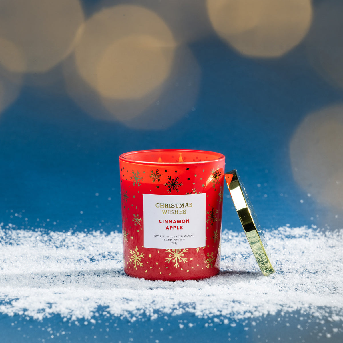 Christmas Wishes Cinnamon Apple 283g scented candle in red glass, lit and displayed on faux snow against a blue festive background with warm blurred lights.