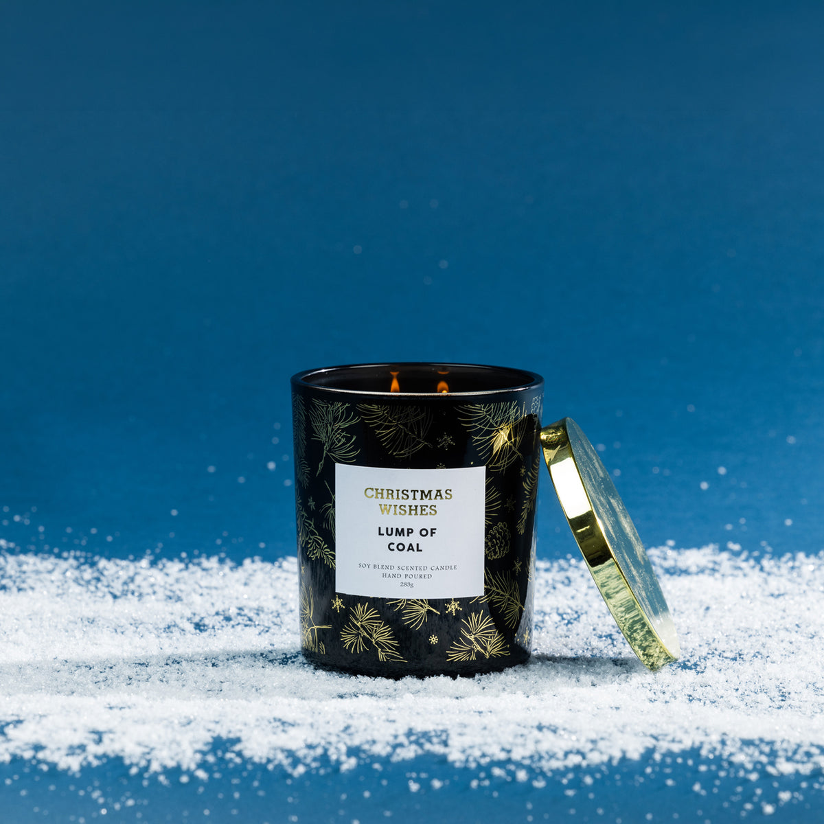 Christmas Wishes Lump of Coal 283g scented candle in a black glass vessel with gold botanical detailing, lit with a gold lid resting beside it.