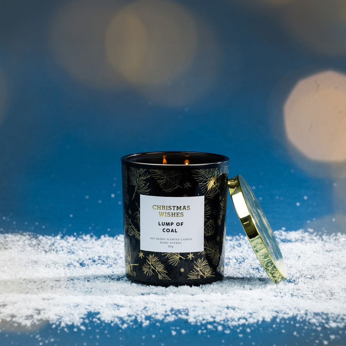 Christmas Wishes Lump of Coal 283g scented candle in black glass, lit and displayed on faux snow against a blue festive background with soft warm lights.