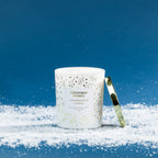 Christmas Wishes Gingerbread Cookie 283g scented candle in a white glass vessel with gold and green botanical details, lit with a gold lid resting beside it.